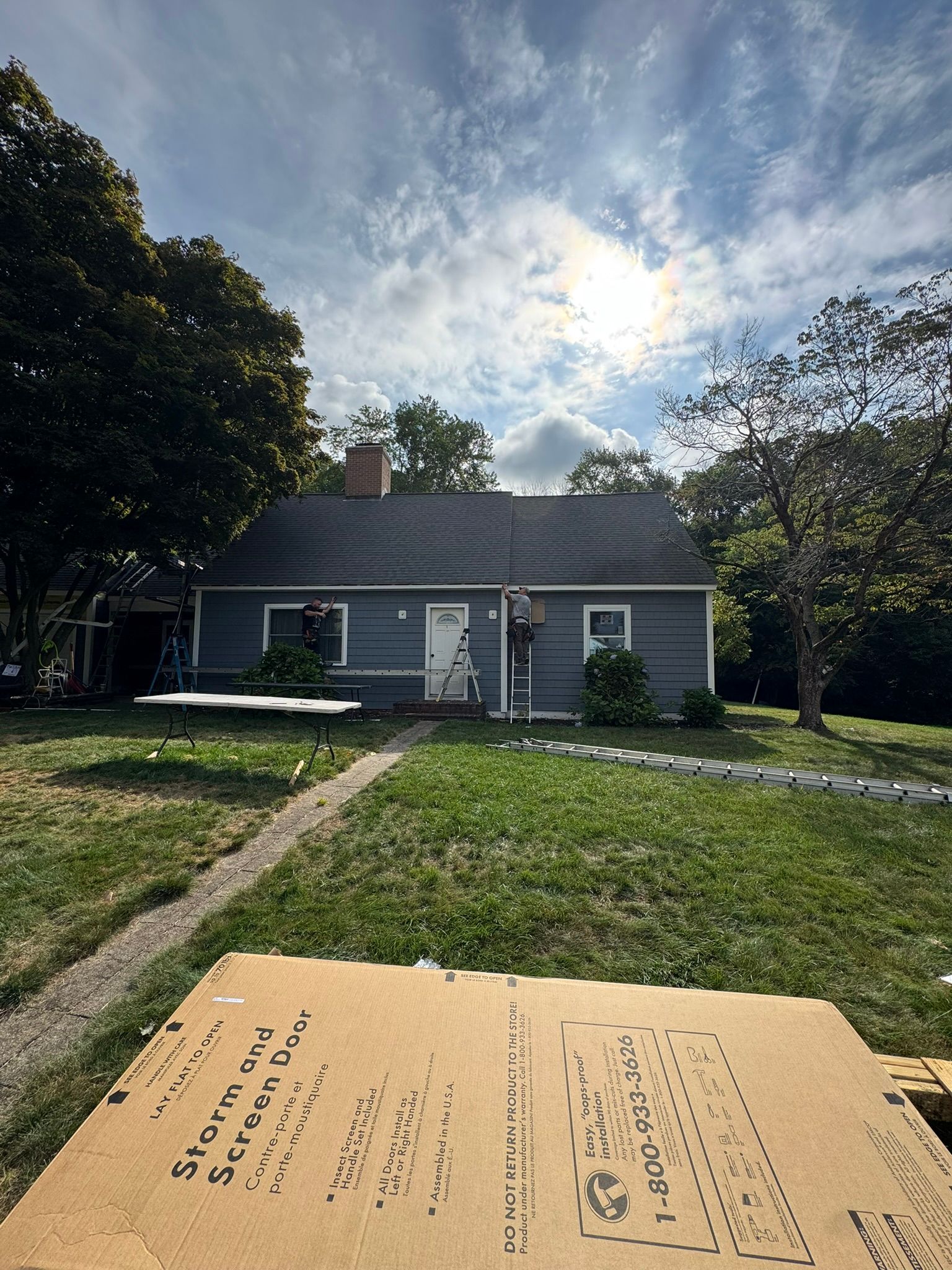 Small gray house with a front lawn and path, seen from a driveway under a bright cloudy sky