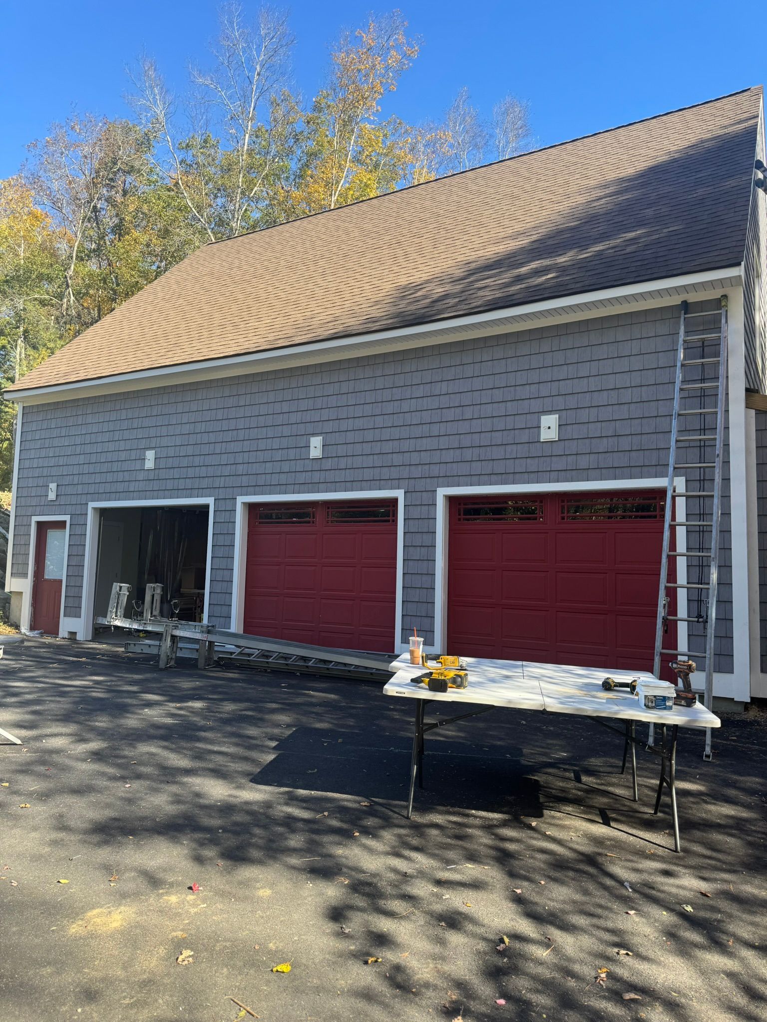 Gray garage building with three red doors and a tan shingled roof, with work tables in front