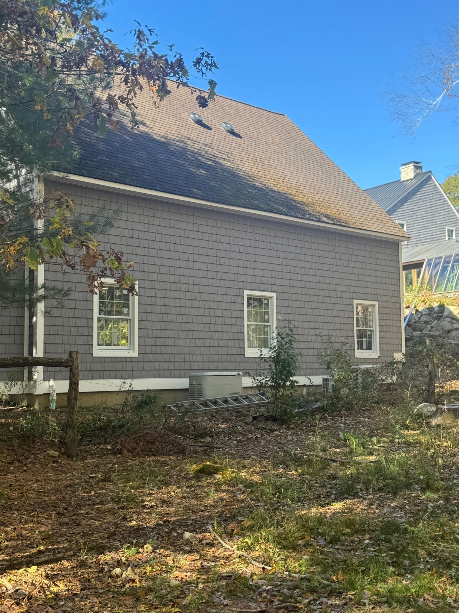 Gray house with a steep roof and white-trimmed windows, partly shaded by trees in a leaf-covered yard