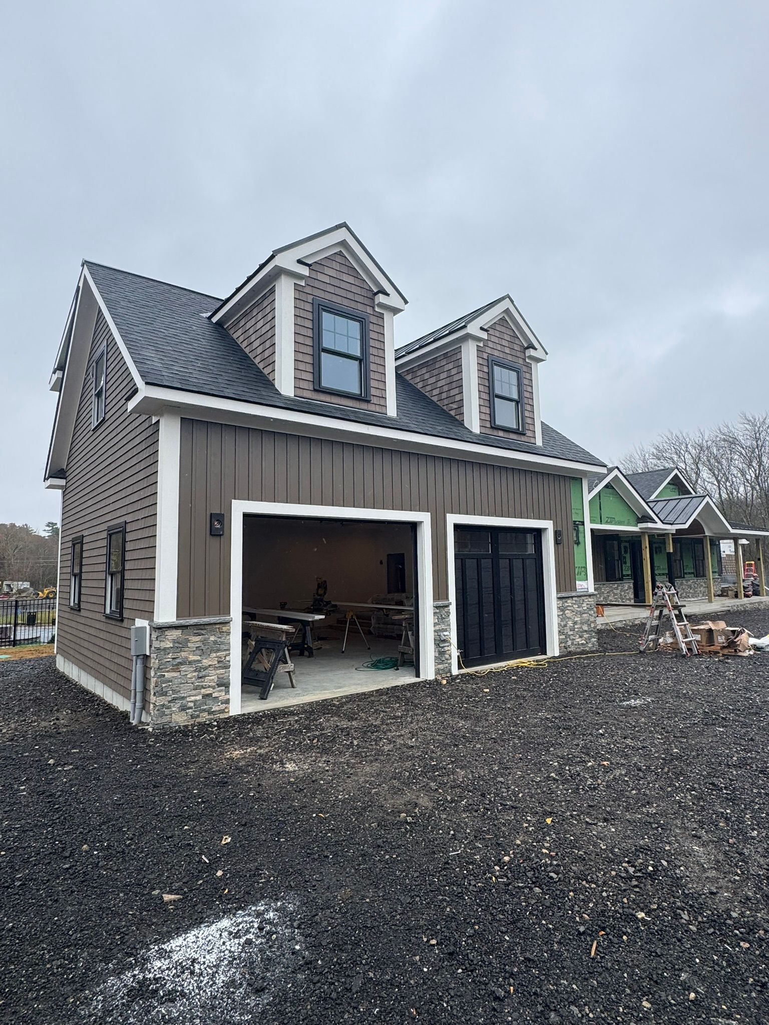 New house under construction with stone siding, dark roof, and open garage on a gravel lot