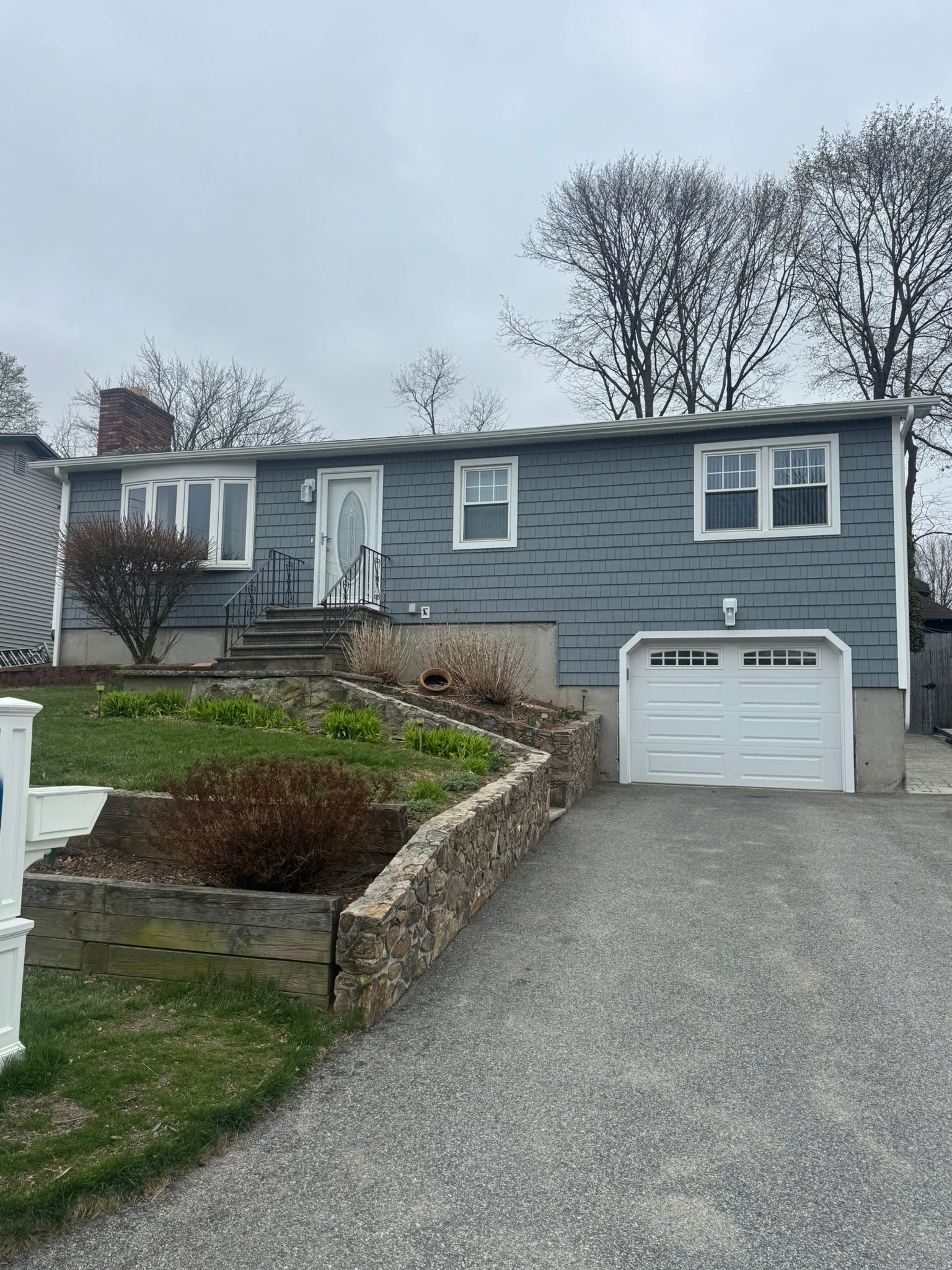 Blue house with a white garage on a sloped driveway, stone retaining wall, and bare trees behind it