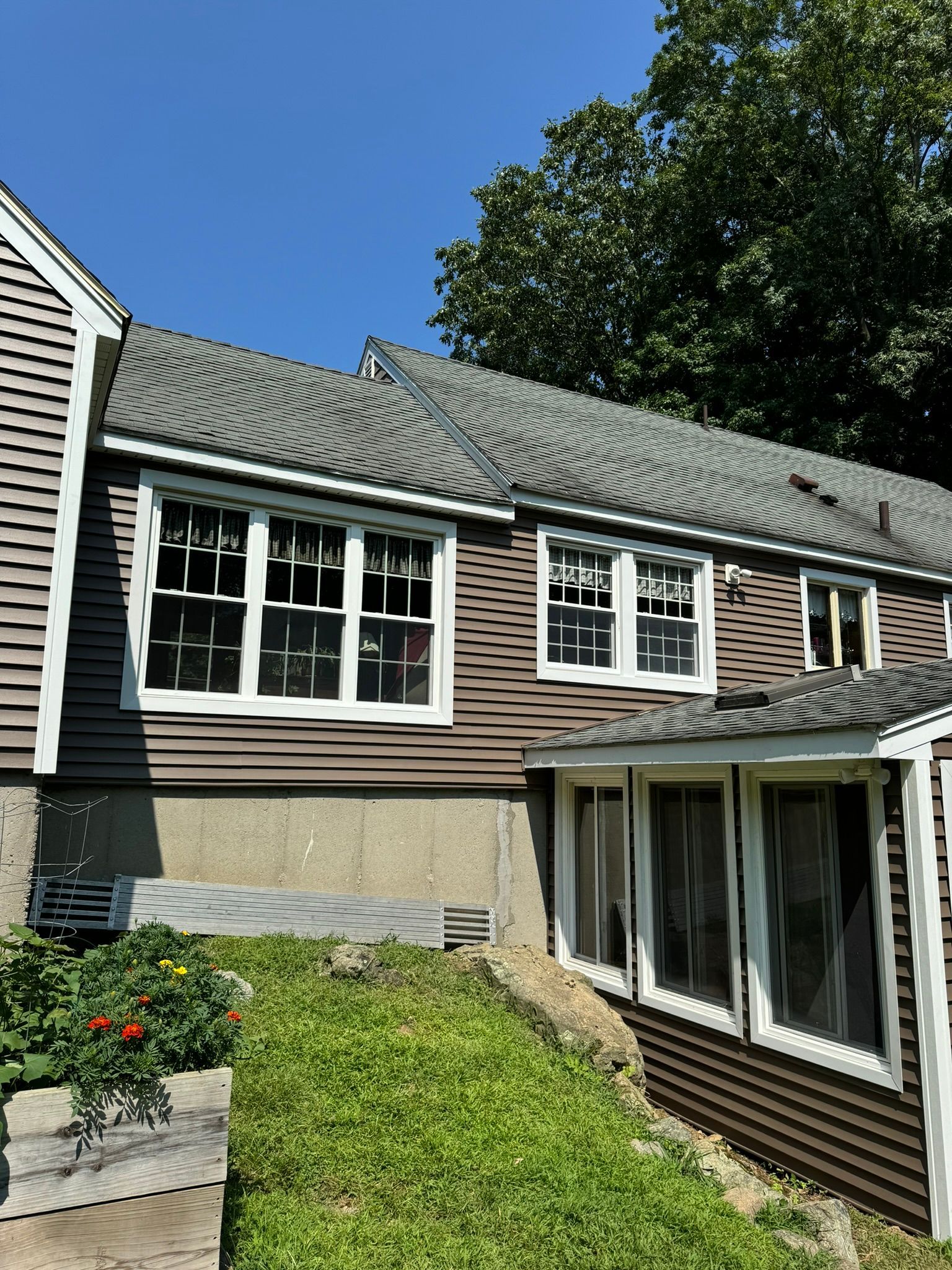 Two-story house exterior with gray roof, beige siding, white-trimmed windows, and a small garden slope