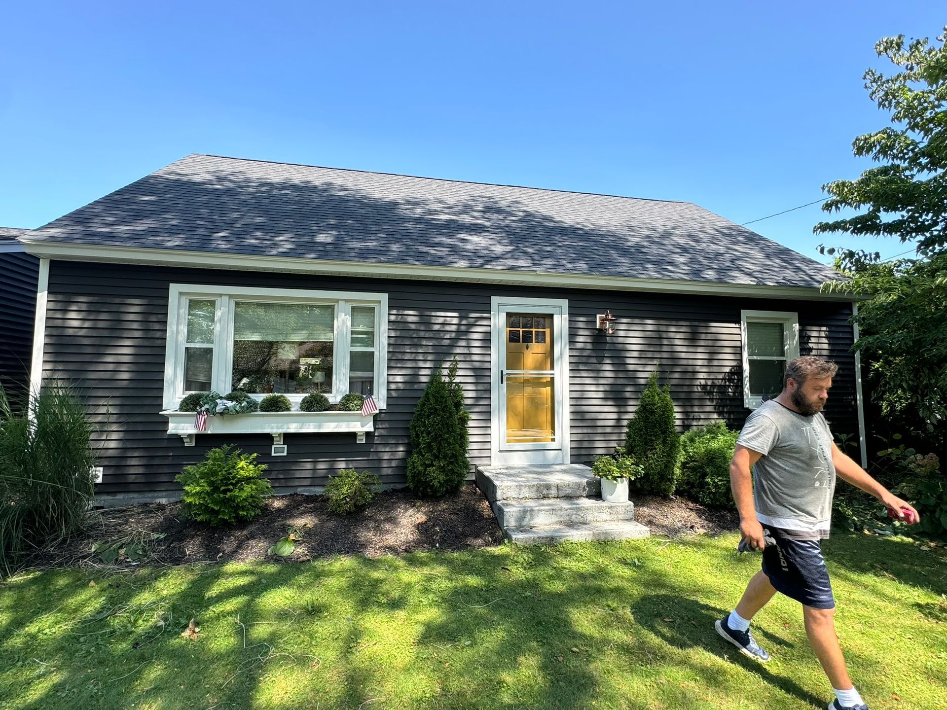 Small dark house with a front yard; a person walks across the lawn under a blue sky.