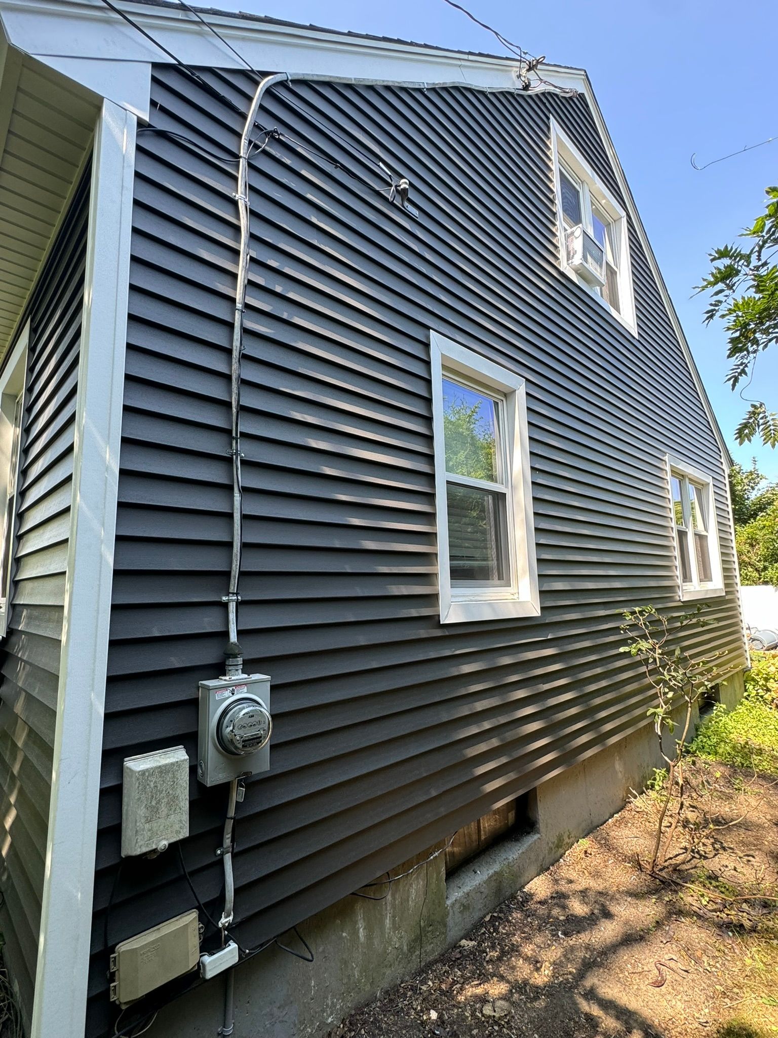 Side of a house with dark gray siding, windows, utility meters, and a concrete foundation under a blue sky