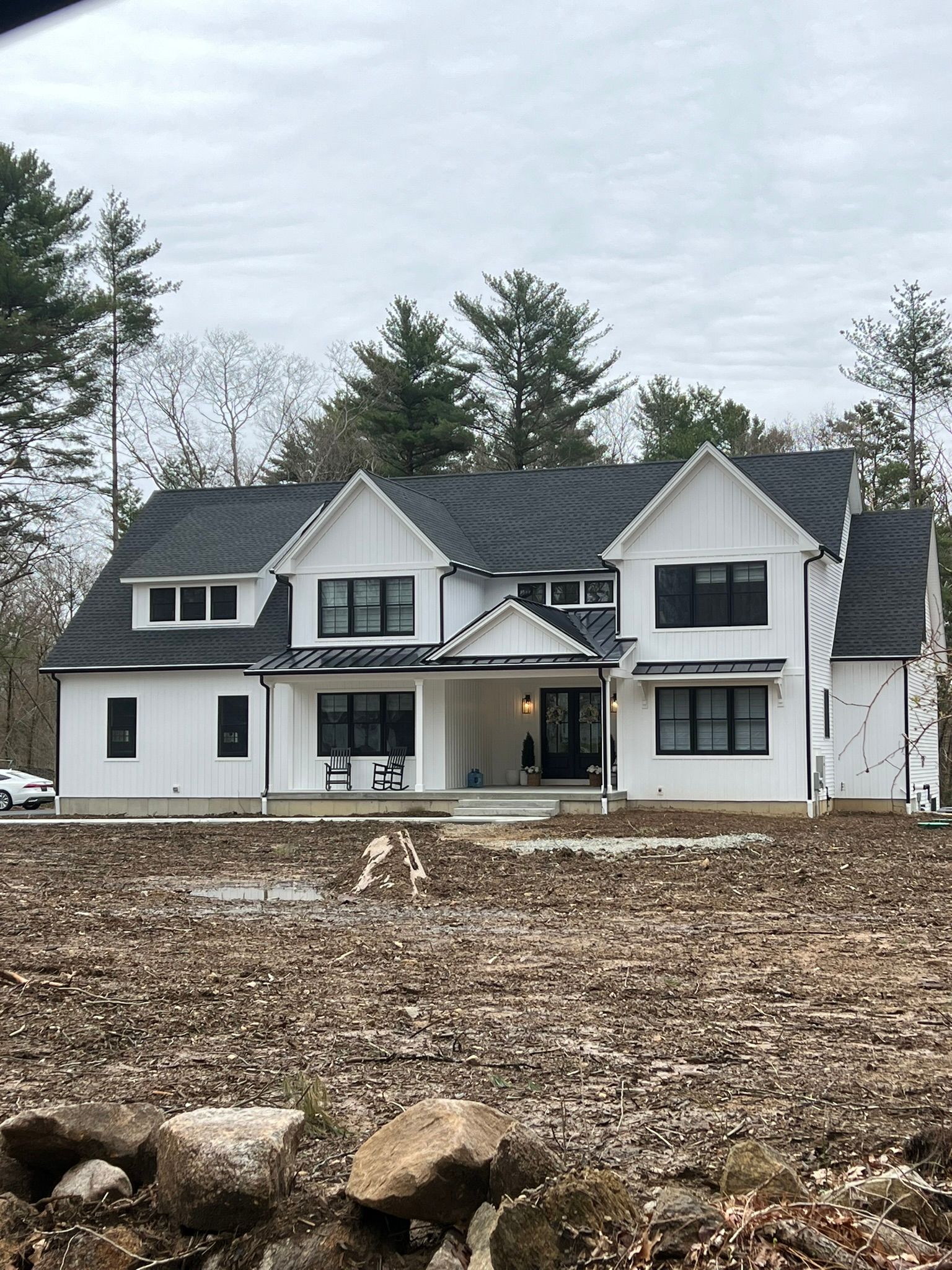 White two-story house under construction with dark roof, bare yard, and trees in the background