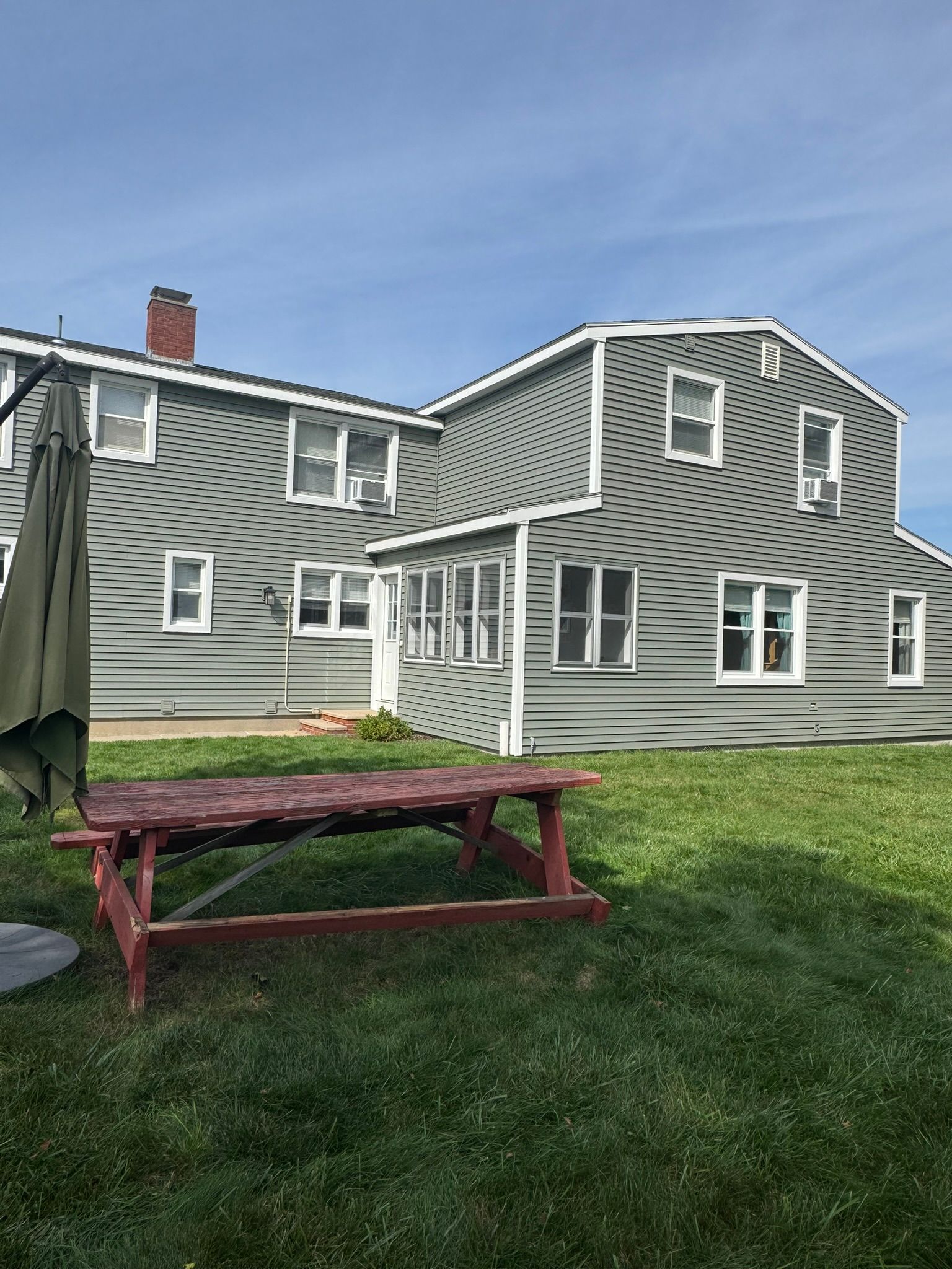 Gray two-story house with white trim and a red picnic table on a green lawn under a blue sky
