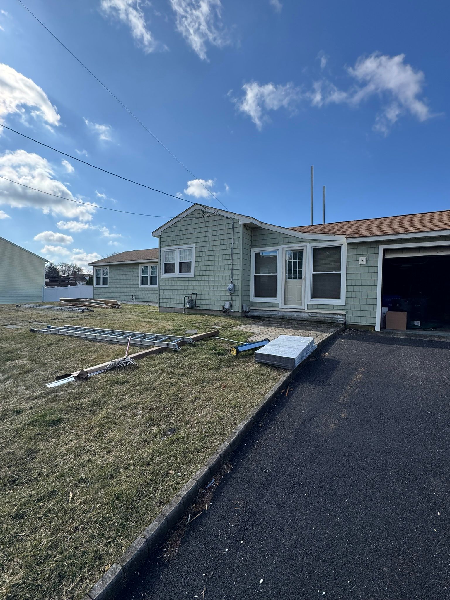 Severely damaged house with broken siding, debris in the yard, and a partly open garage under a blue sky
