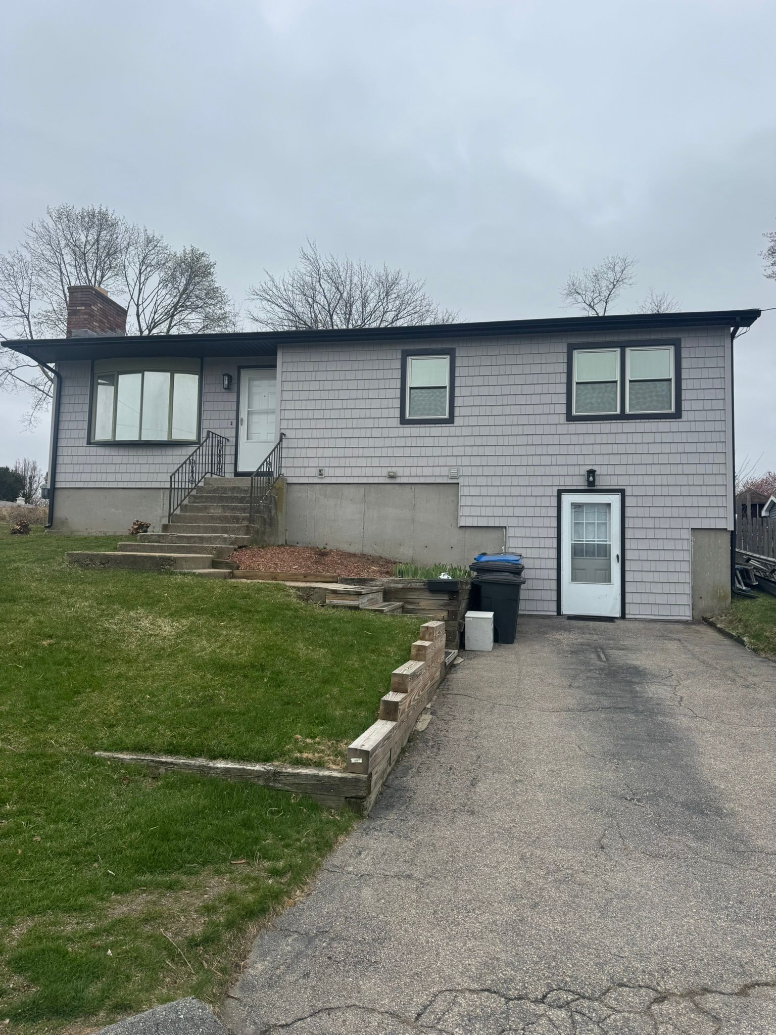 Gray house with front stairs, sloped lawn, and driveway under an overcast sky