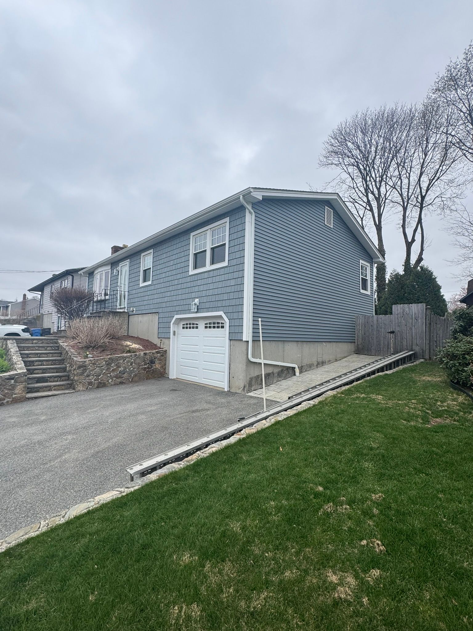 Gray house with curved siding, gravel driveway, white garage, and green lawn on a cloudy day
