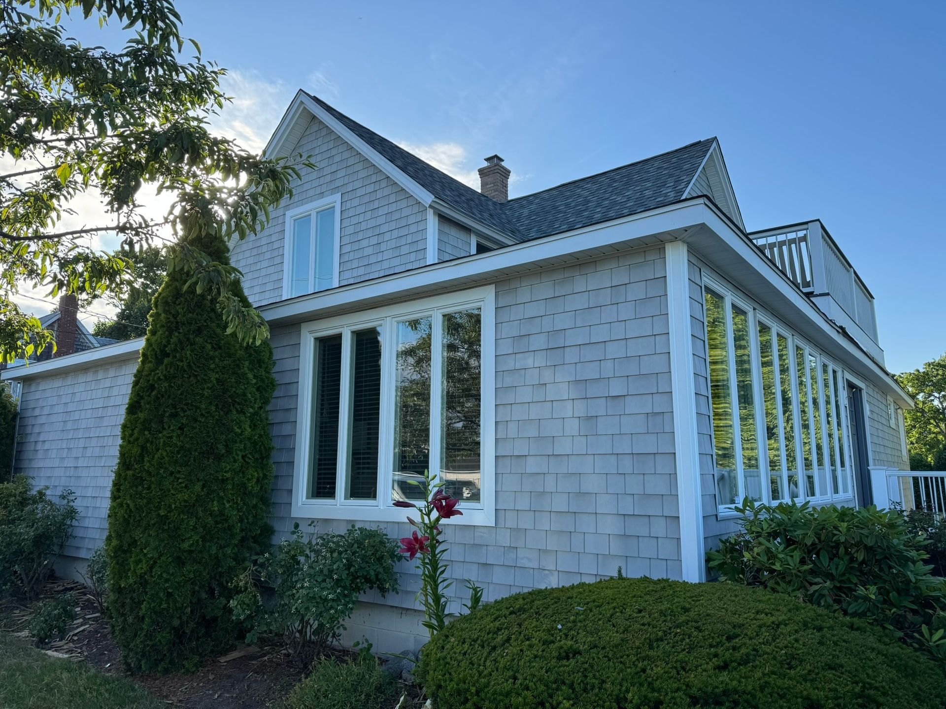 White house with gray shingle siding, large windows, and green shrubs in a sunny garden.