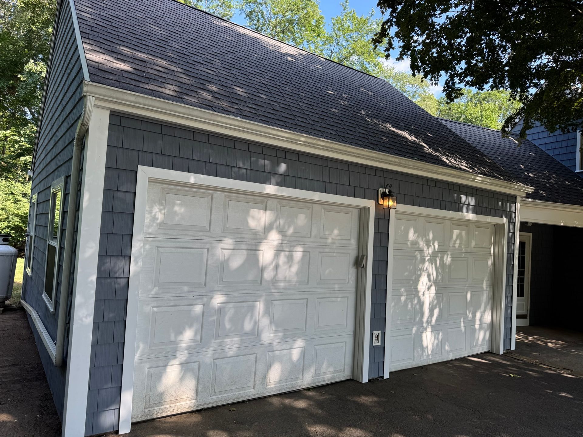 Two white garage doors on a gray detached garage with shingled roof and trees overhead