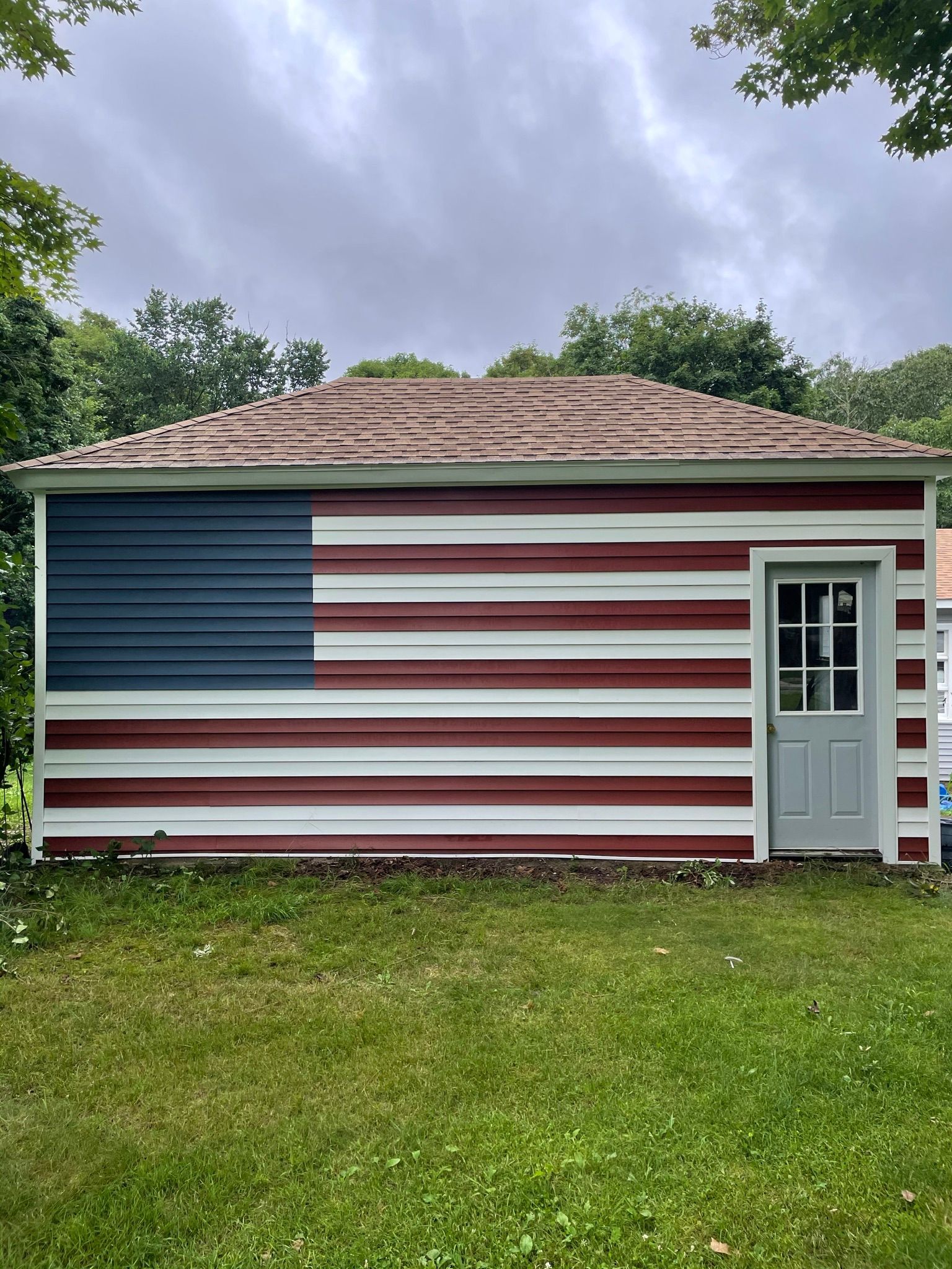 Small shed painted like the U.S. flag, with a gray door, in a grassy yard under cloudy sky