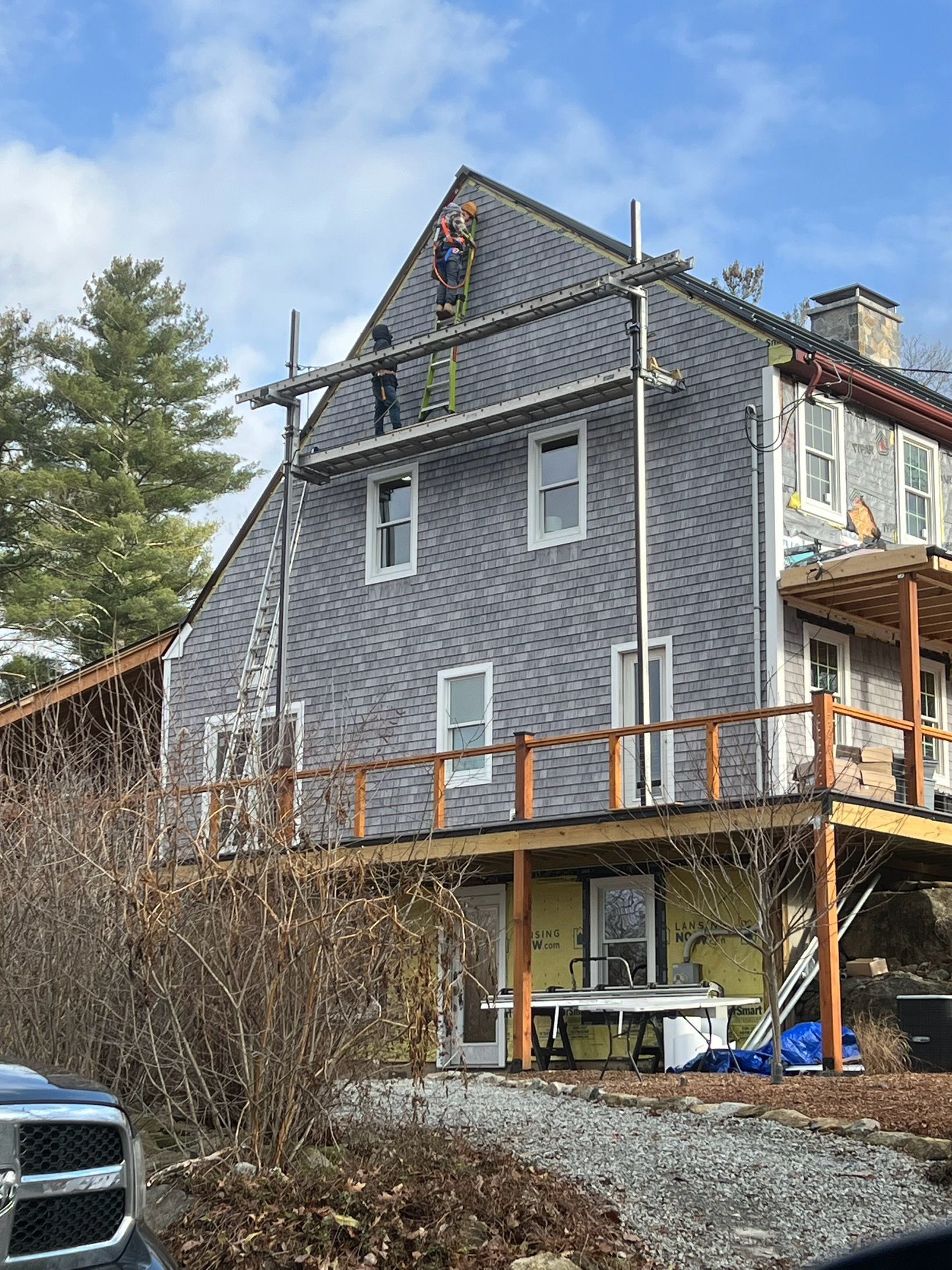 Modern house under construction with gray exterior siding, wooden deck, and trees in the background
