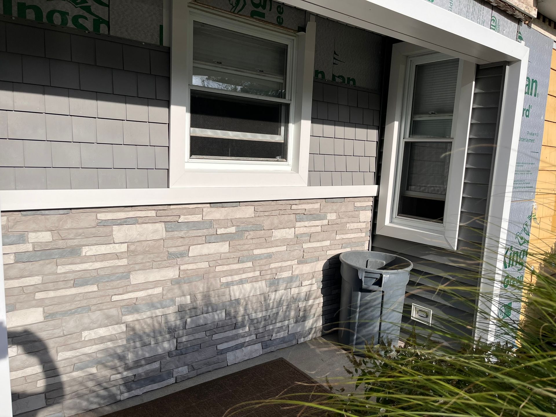 House exterior with gray siding, stone facade, white windows, and a small trash bin near the porch.