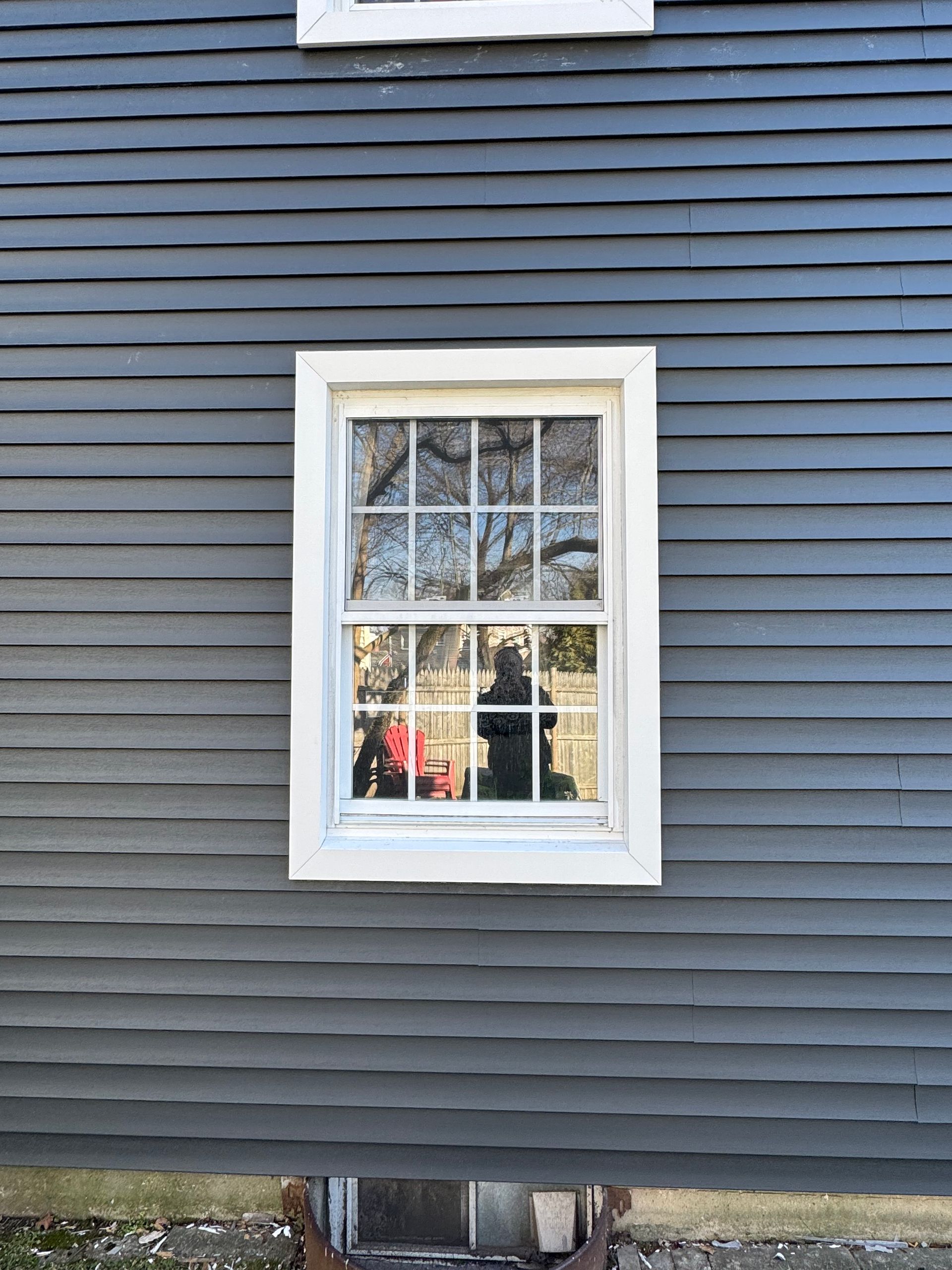 White-framed window on blue-gray siding, reflecting trees and a red object inside.
