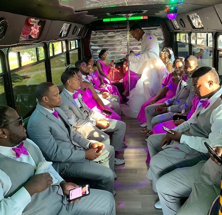 Wedding party in a bus; bride in white dress, bridesmaids in pink, groomsmen in gray, all looking at something.
