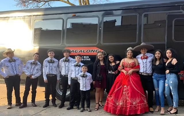 Group posing in front of a black bus. Includes a mariachi band, a girl in a red dress, and friends/family.