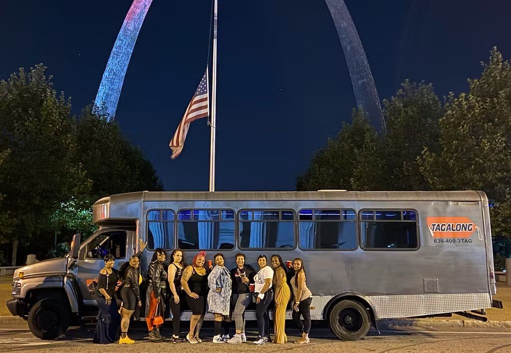 People pose by a party bus with the St. Louis Arch in the background at night.