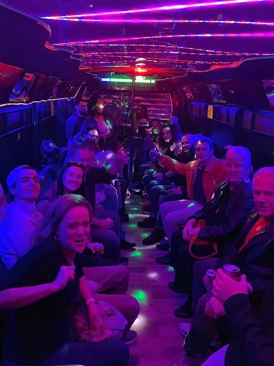 People partying on a party bus with neon lights. Passengers are sitting, some are smiling and waving.
