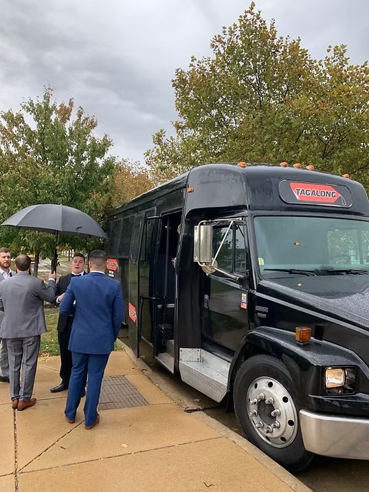 Group of men in suits boarding a black shuttle bus, one holding an umbrella on a cloudy day.