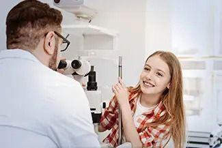 A person smiles while sitting at an eye examination machine, looking toward the doctor operating the equipment.