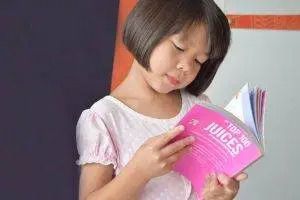 A person with short dark hair wearing a light pink top, focused on reading a bright pink book titled 