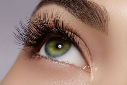 A close-up of a human eye with green iris, long dark eyelashes, and groomed eyebrows against a soft-focus background.