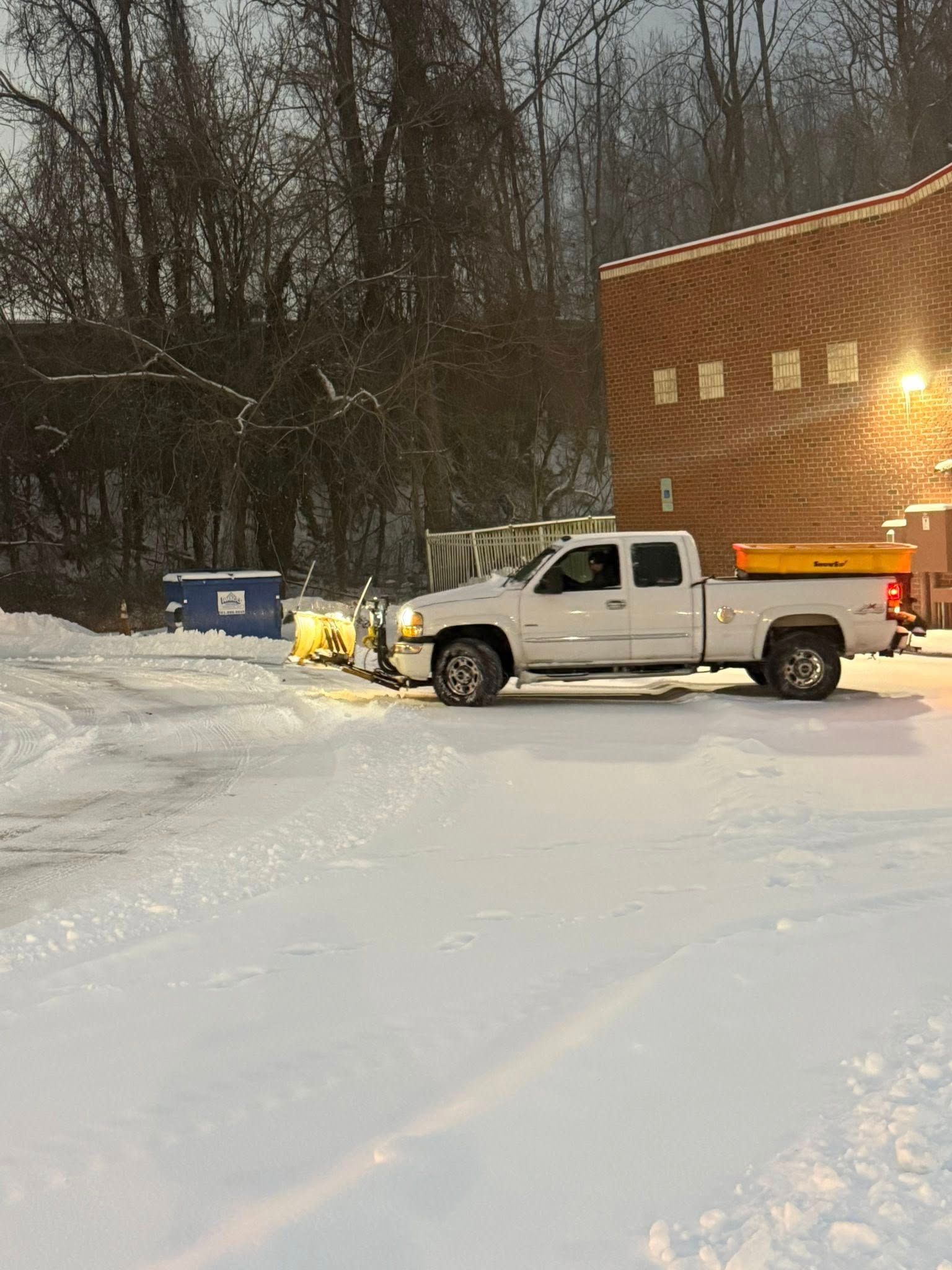 A white pickup truck with a yellow snowplow attached in a snowy parking lot next to a brick building.