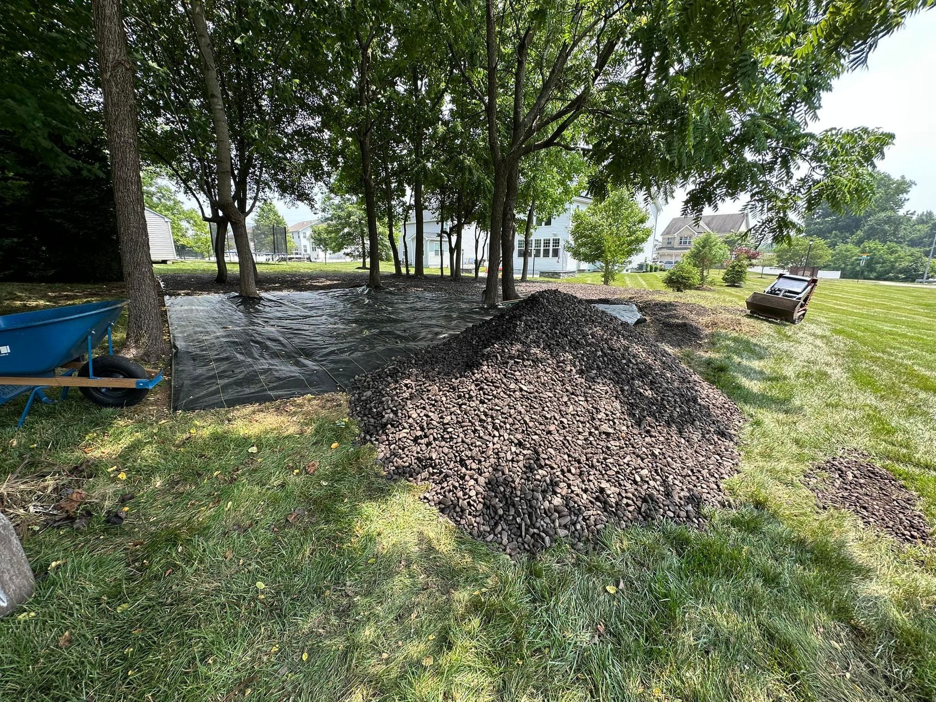 A large pile of dark mulch sits next to a patch of black landscaping fabric laid out beneath trees in a grassy yard.