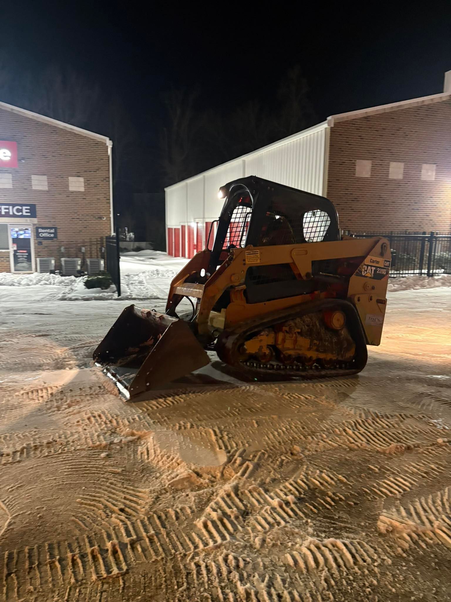 A yellow skid-steer loader parked on snow-covered ground at night, with industrial buildings in the background.