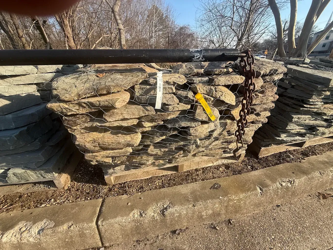 A pallet of stacked, irregularly shaped brown and grey natural stones outdoors, secured with a chain and a black metal bar.