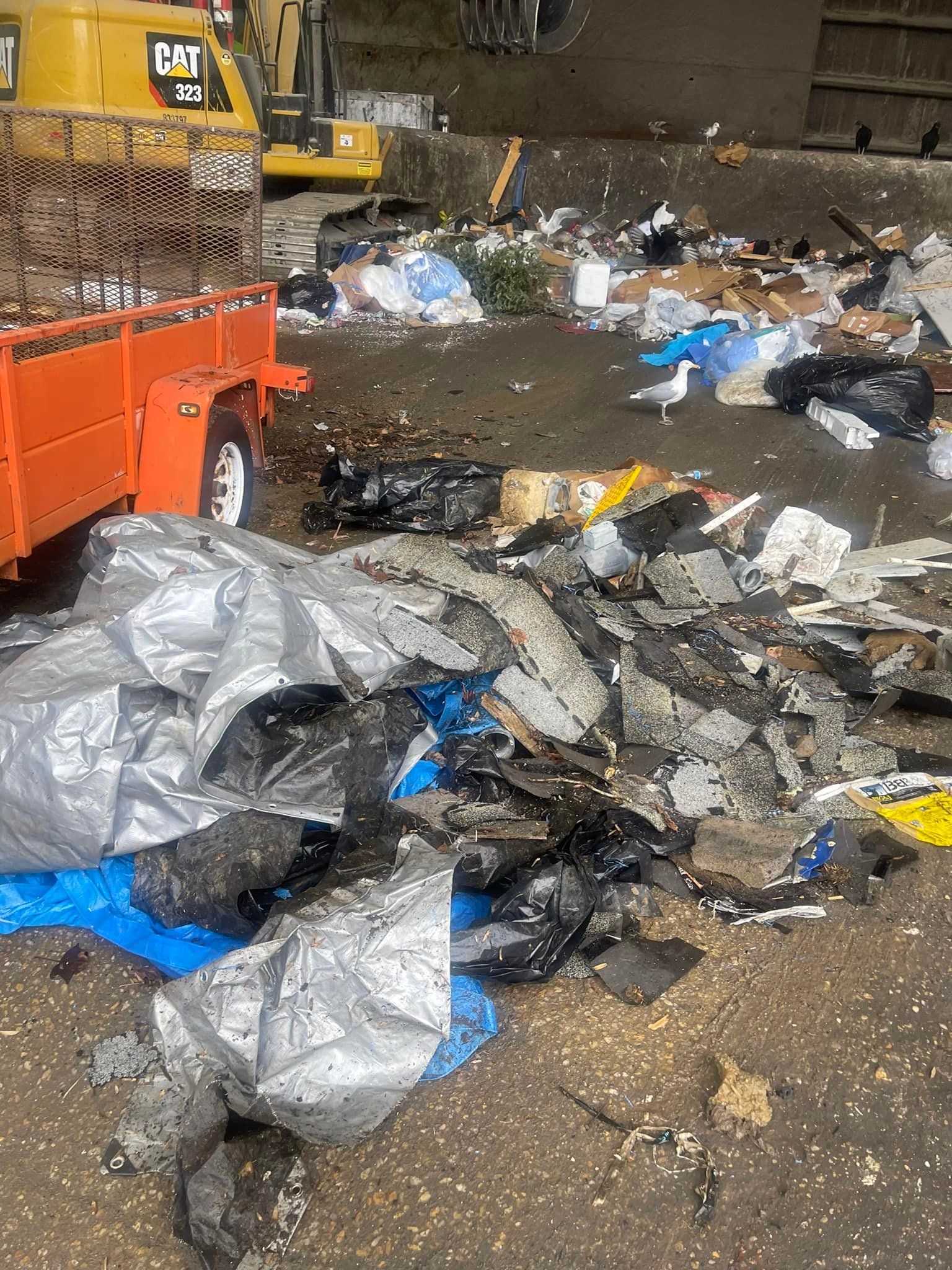 A debris-strewn industrial site features a yellow CAT excavator, an orange trailer, and scattered refuse on the ground.