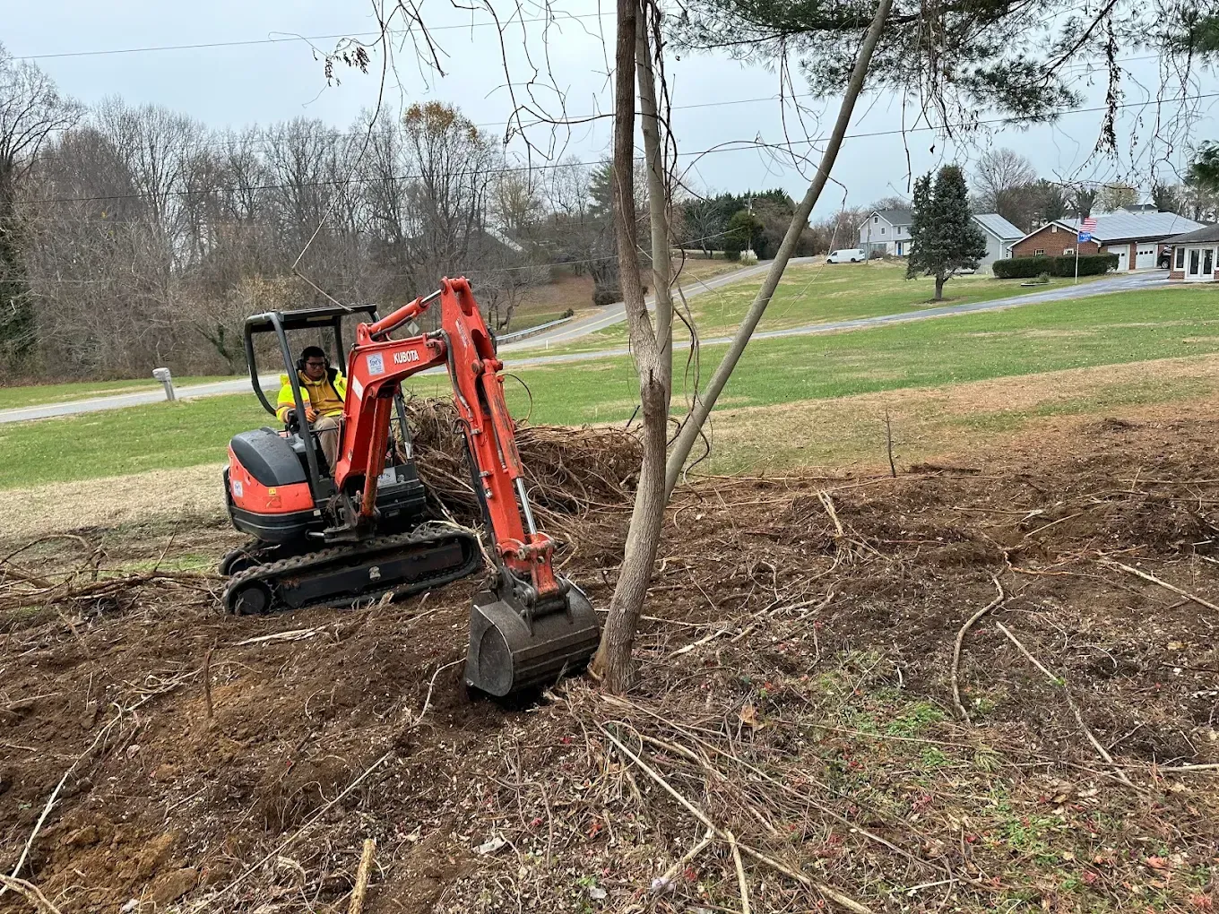 An orange compact excavator sits on a dirt-covered slope in a field, clearing away brush and vegetation.