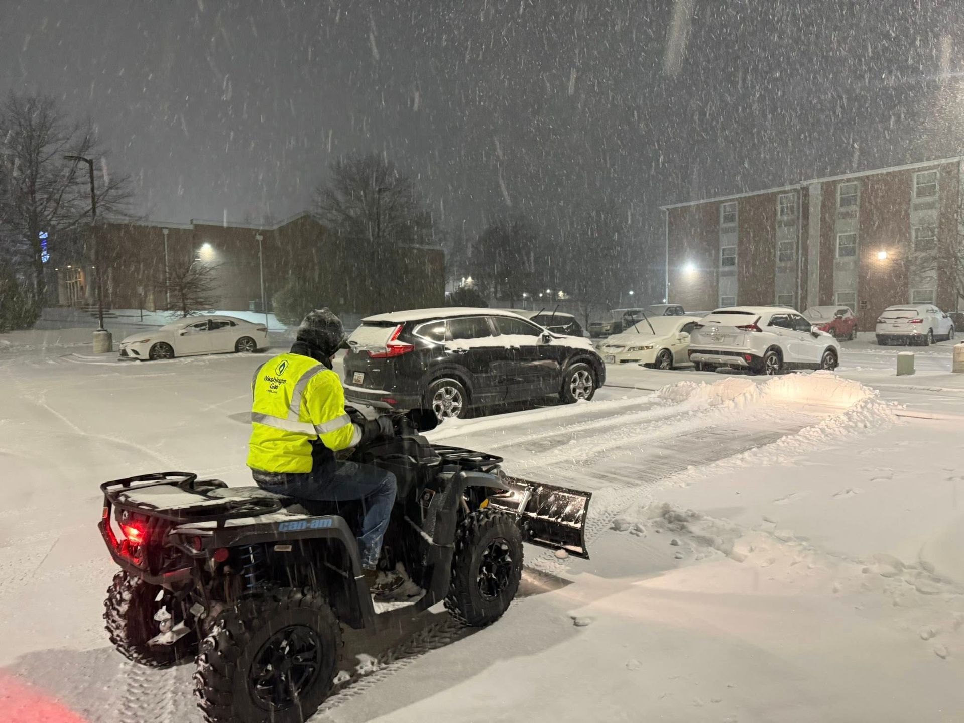 A person wearing a high-visibility jacket drives an ATV with a snowplow through a snowy parking lot at night.