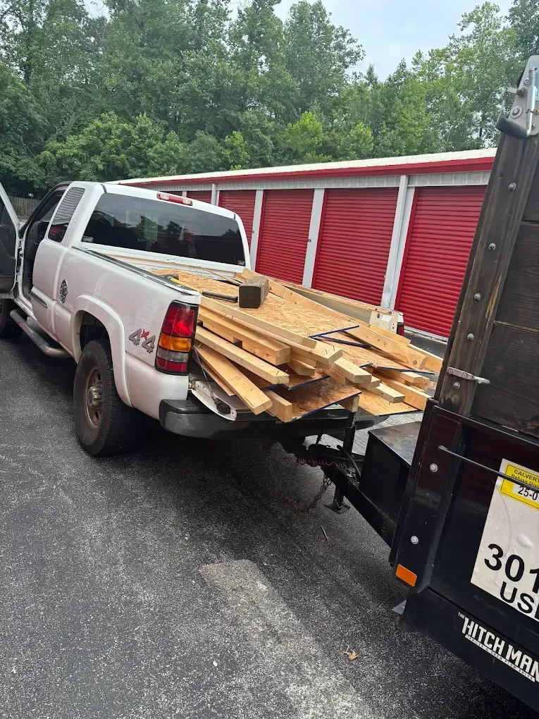 White pickup truck loaded with lumber, hitched to a trailer in front of red and white self-storage units.