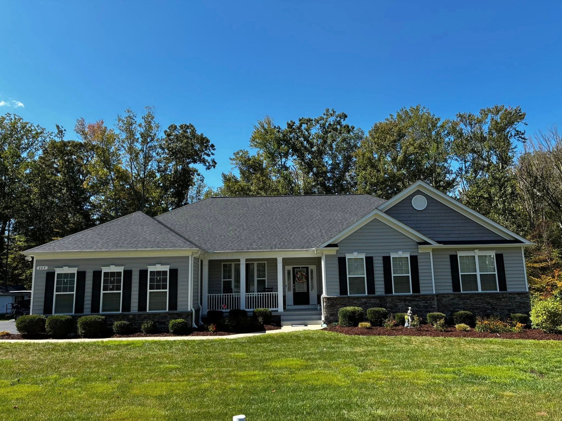 A gray ranch-style house with dark shutters, stone accents, and a dark shingled roof sits before a backdrop of green trees.