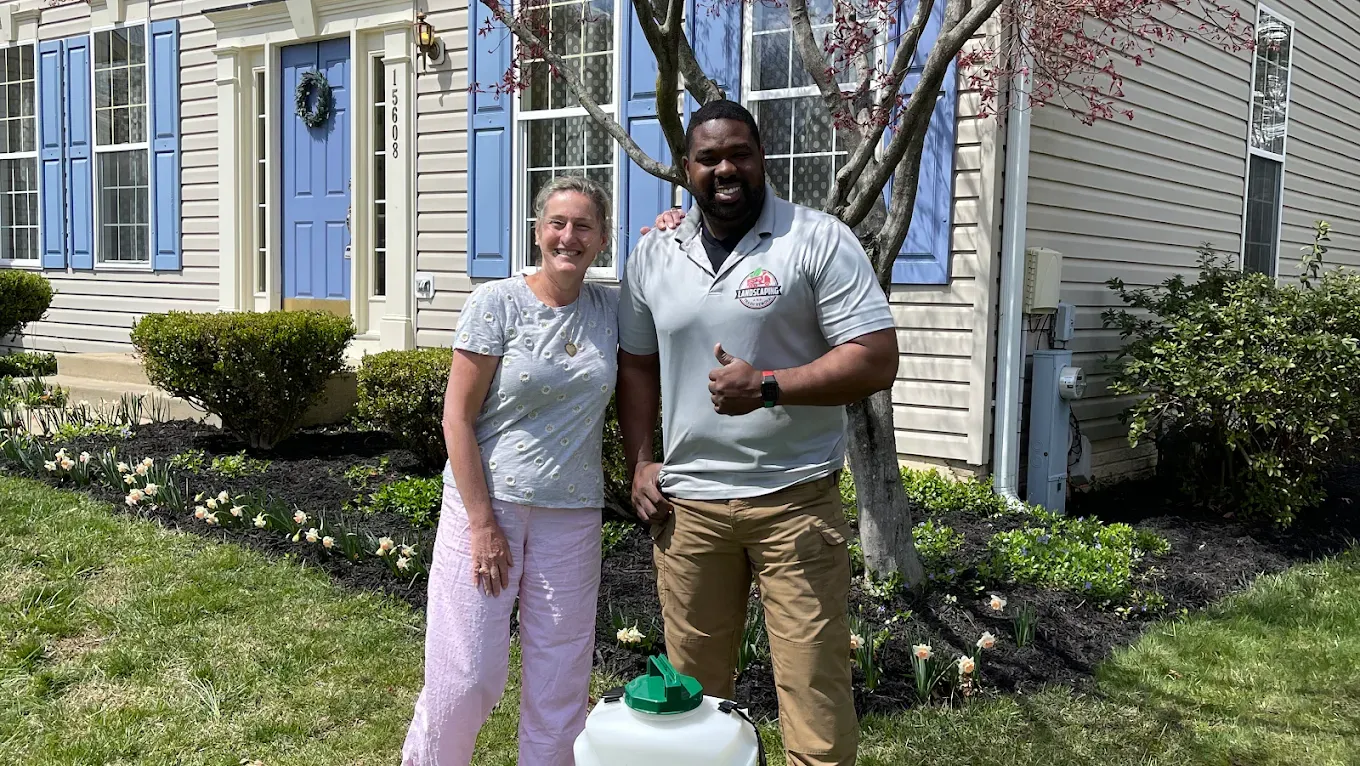 A person and a service technician with a sprayer stand together in front of a house with blue shutters and a blue door.