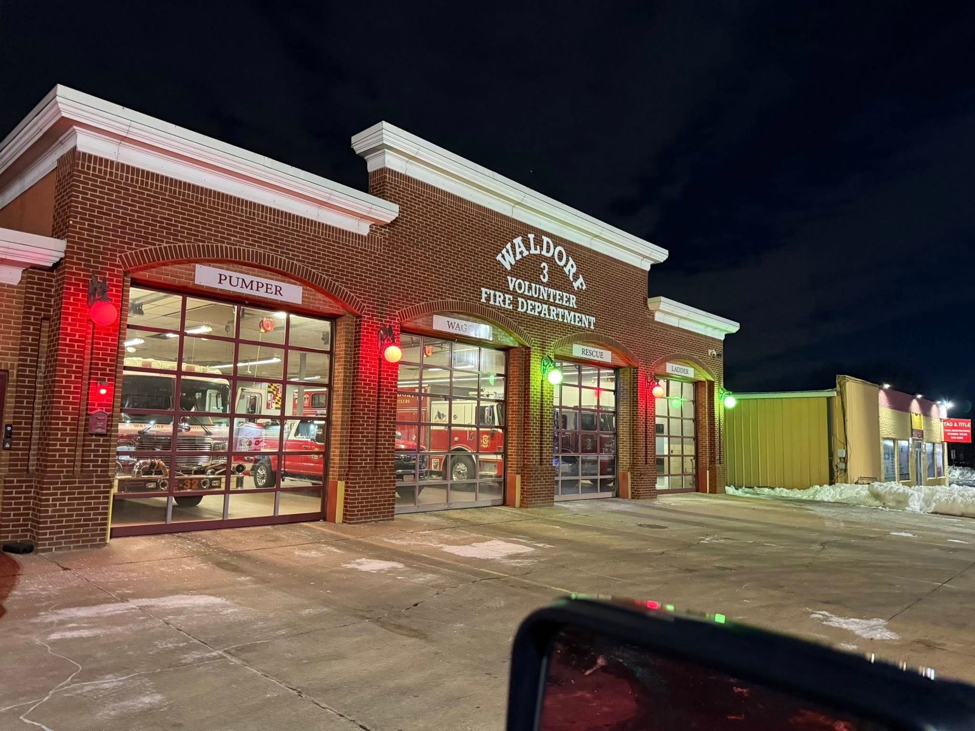 A brick fire station at night with three open garage bays containing fire trucks, viewed from a vehicle.