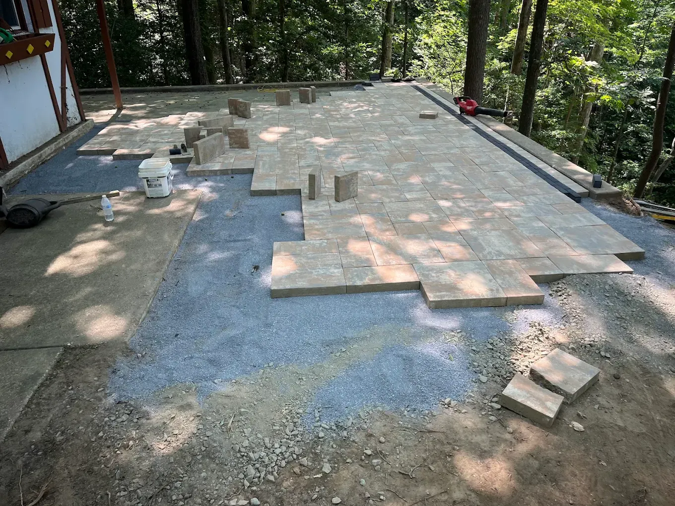 Tan stone pavers being laid on a gravel bed to construct a patio next to a building in a wooded yard.