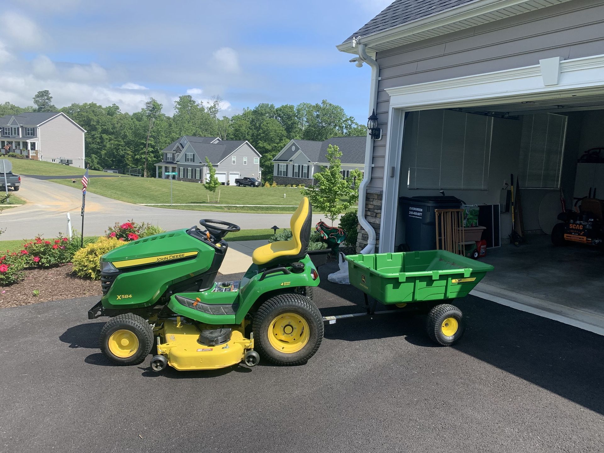 A green John Deere lawn tractor with an attached green utility cart parked on a paved driveway in front of a garage.