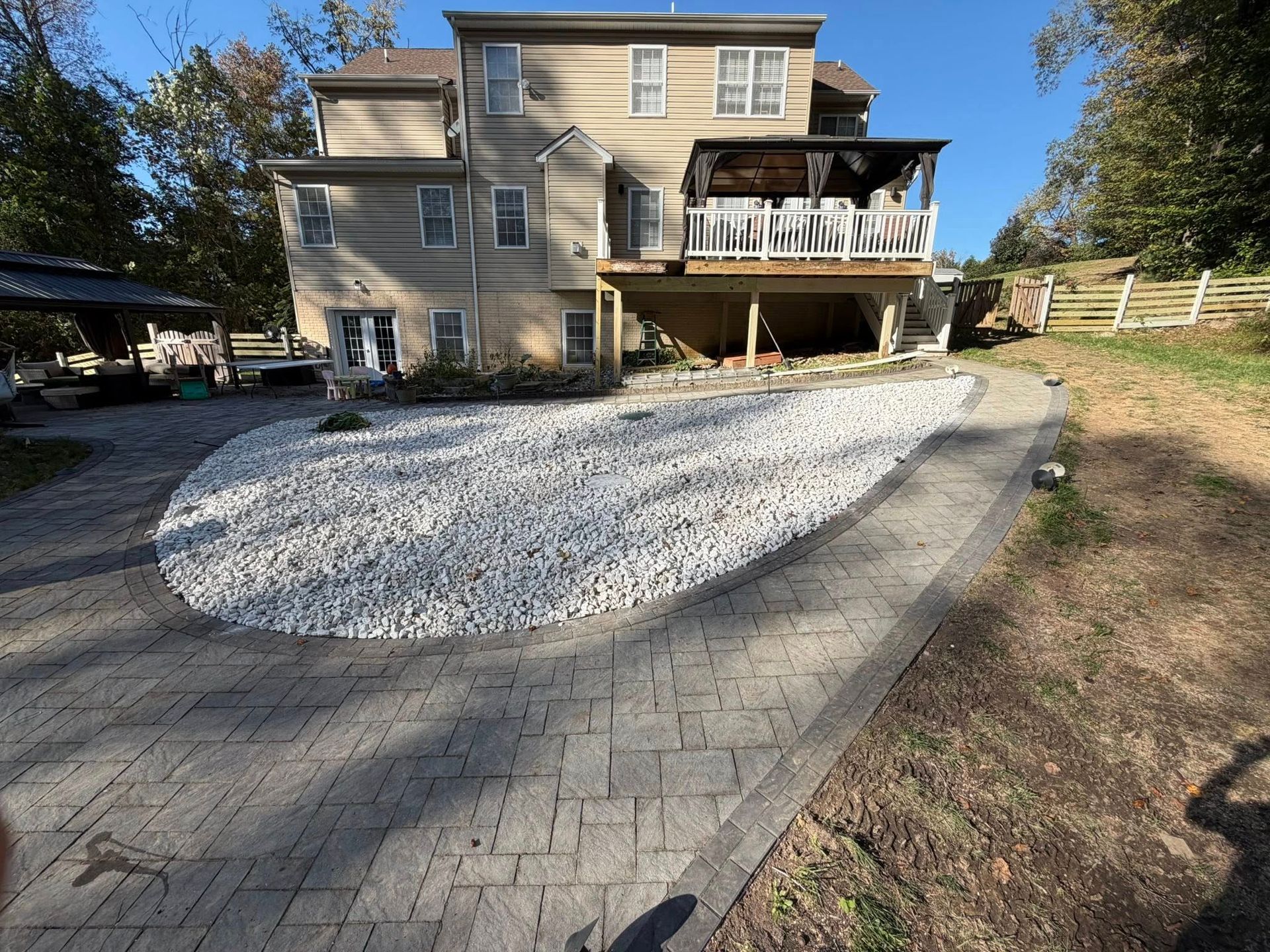 A multi-story beige house with a stone patio, large white rock bed, and elevated wooden deck under a bright blue sky.