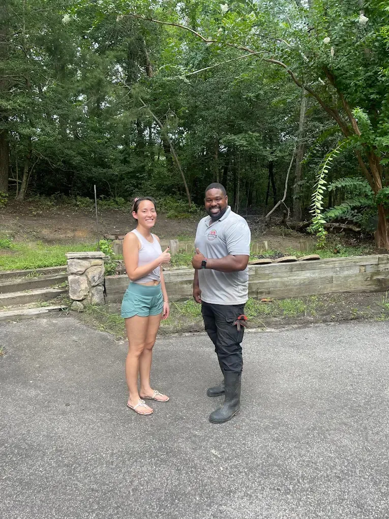 Two people standing on a driveway, smiling and giving a thumbs-up in front of a stone wall and wooded area.