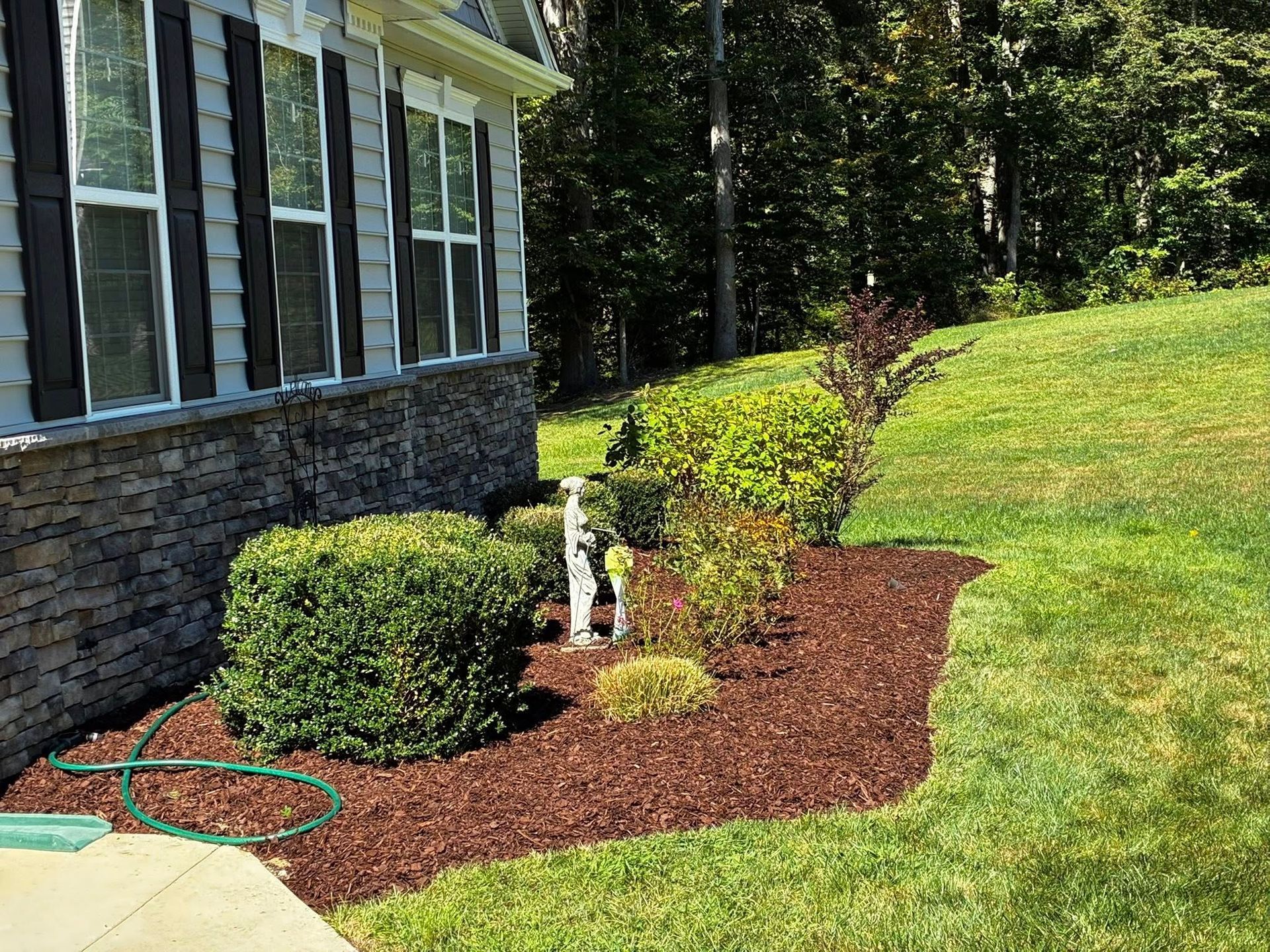 A side view of a stone and siding house with a mulched garden bed containing shrubs and a small statue.