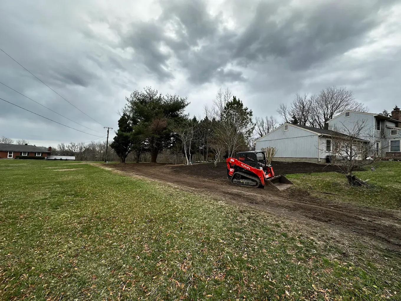 A red skid steer loader performs grading work on a grassy, overcast residential property.