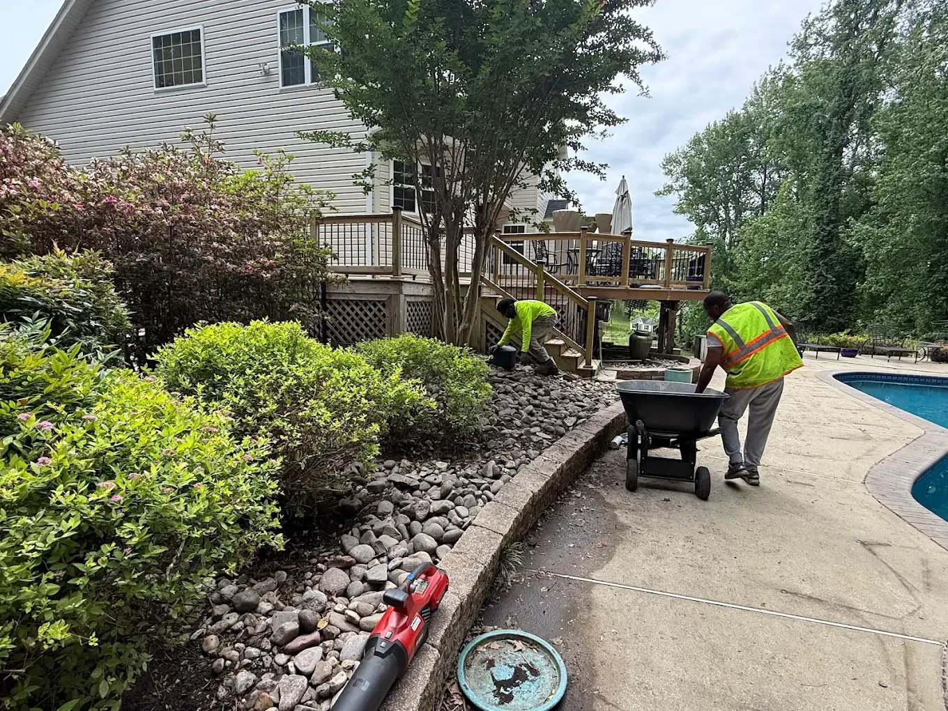 Two workers in neon vests perform landscaping near a house deck and a swimming pool.