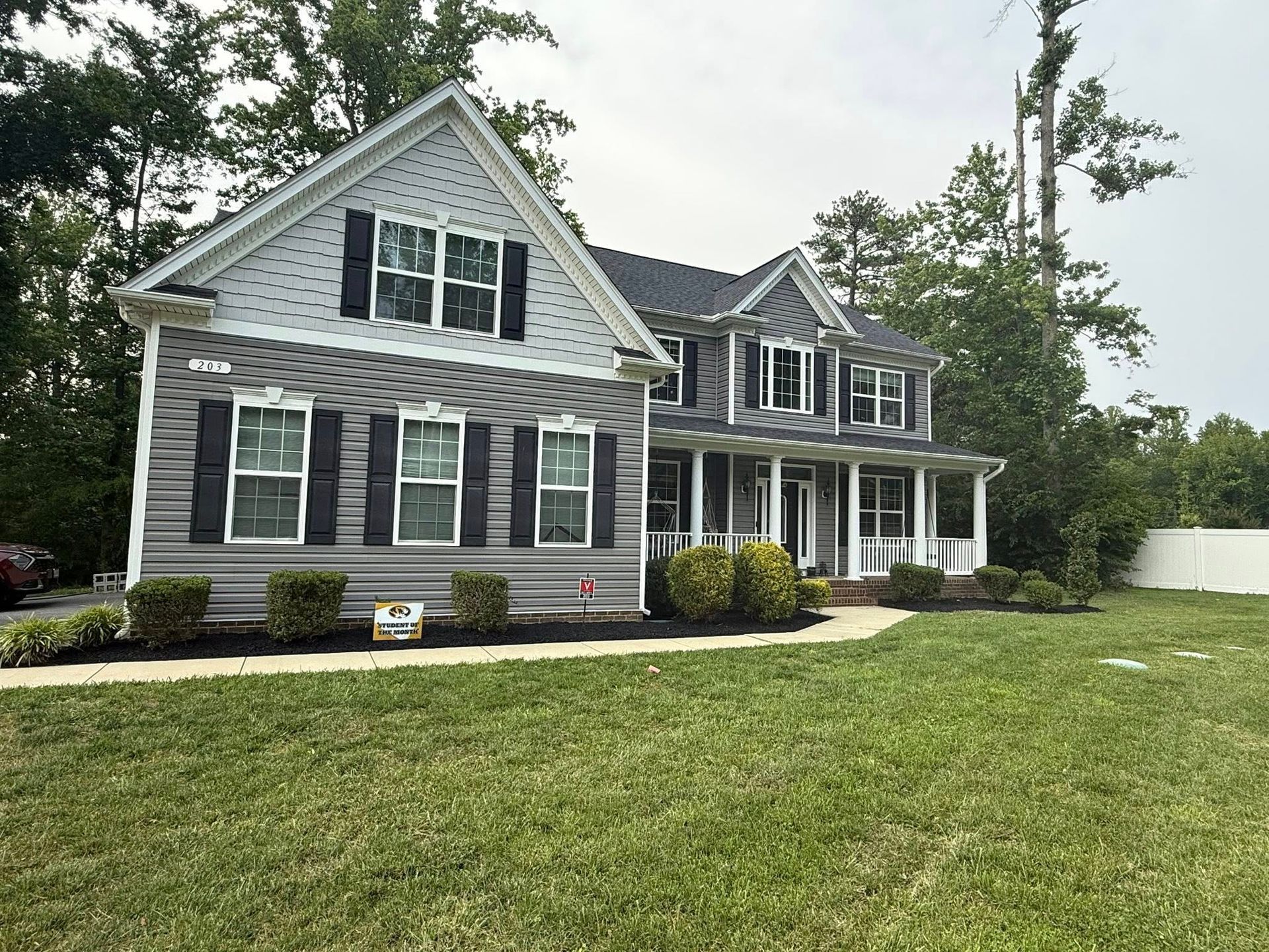A two-story grey suburban house with dark shutters and a front porch, surrounded by green grass and tall trees.