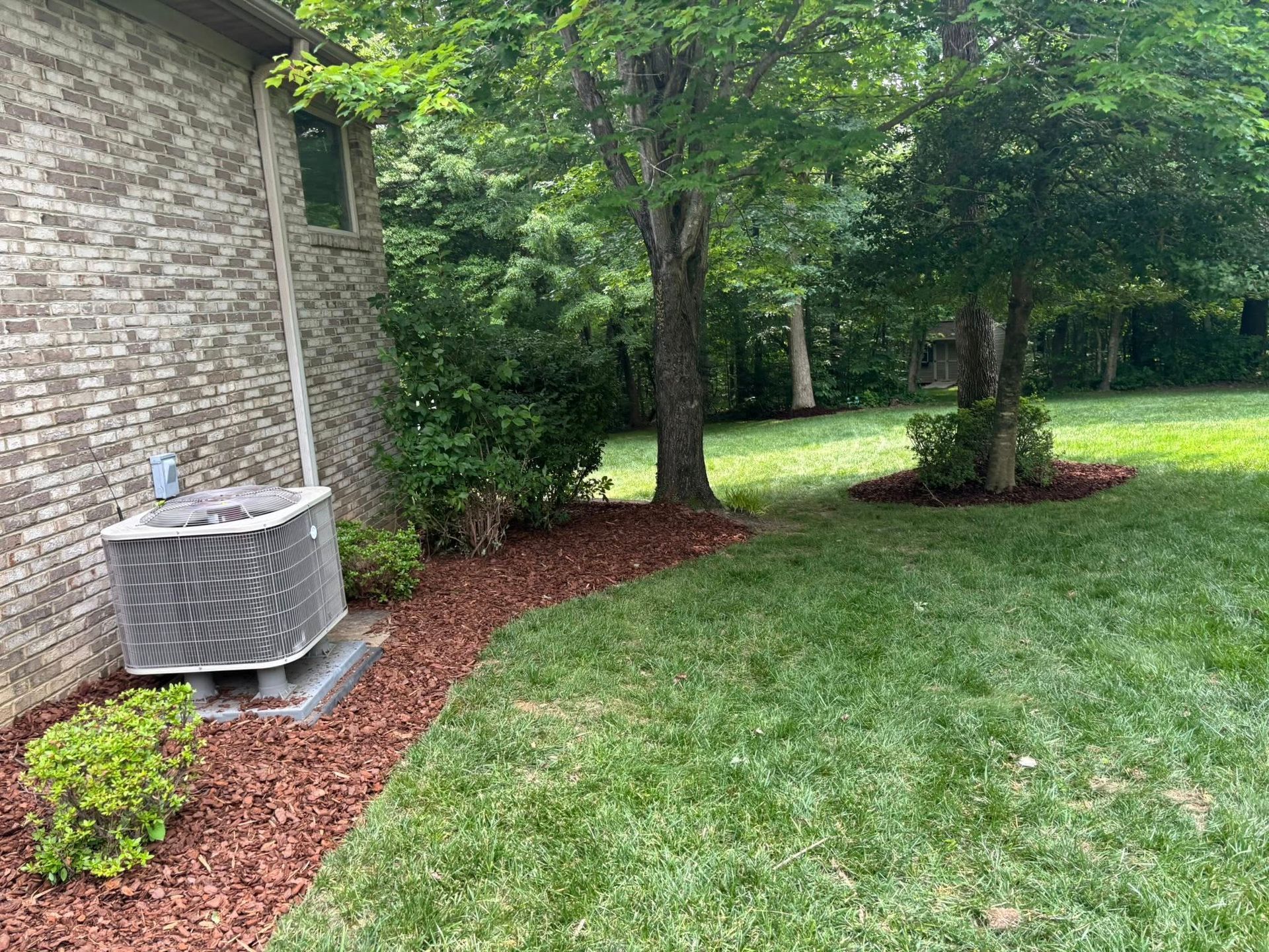 An AC unit sits next to a brick house wall with a mulched garden bed, green lawn, and trees in a backyard.