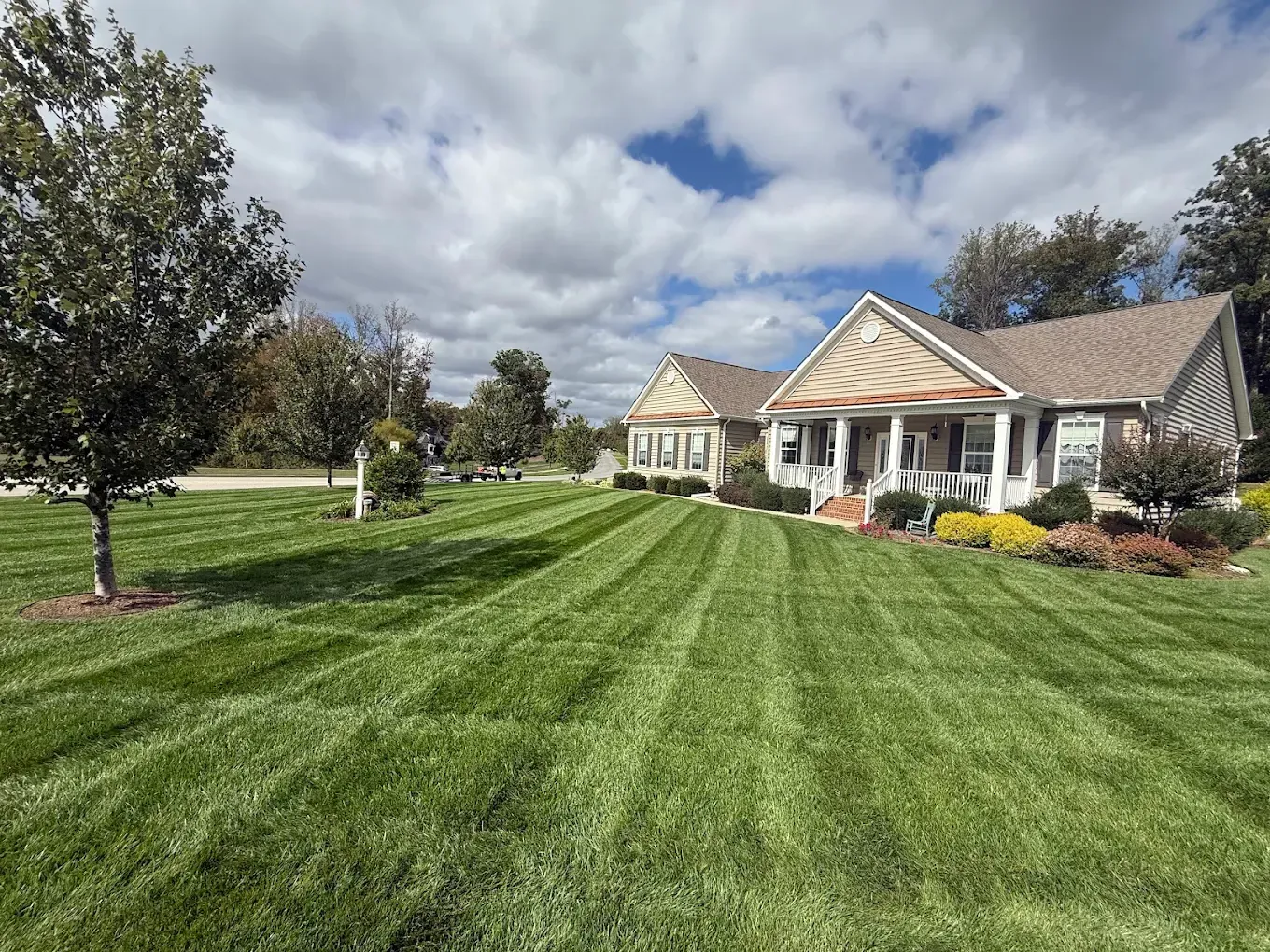 A single-story tan house with a front porch, surrounded by a freshly mowed, striped lawn under a cloudy blue sky.