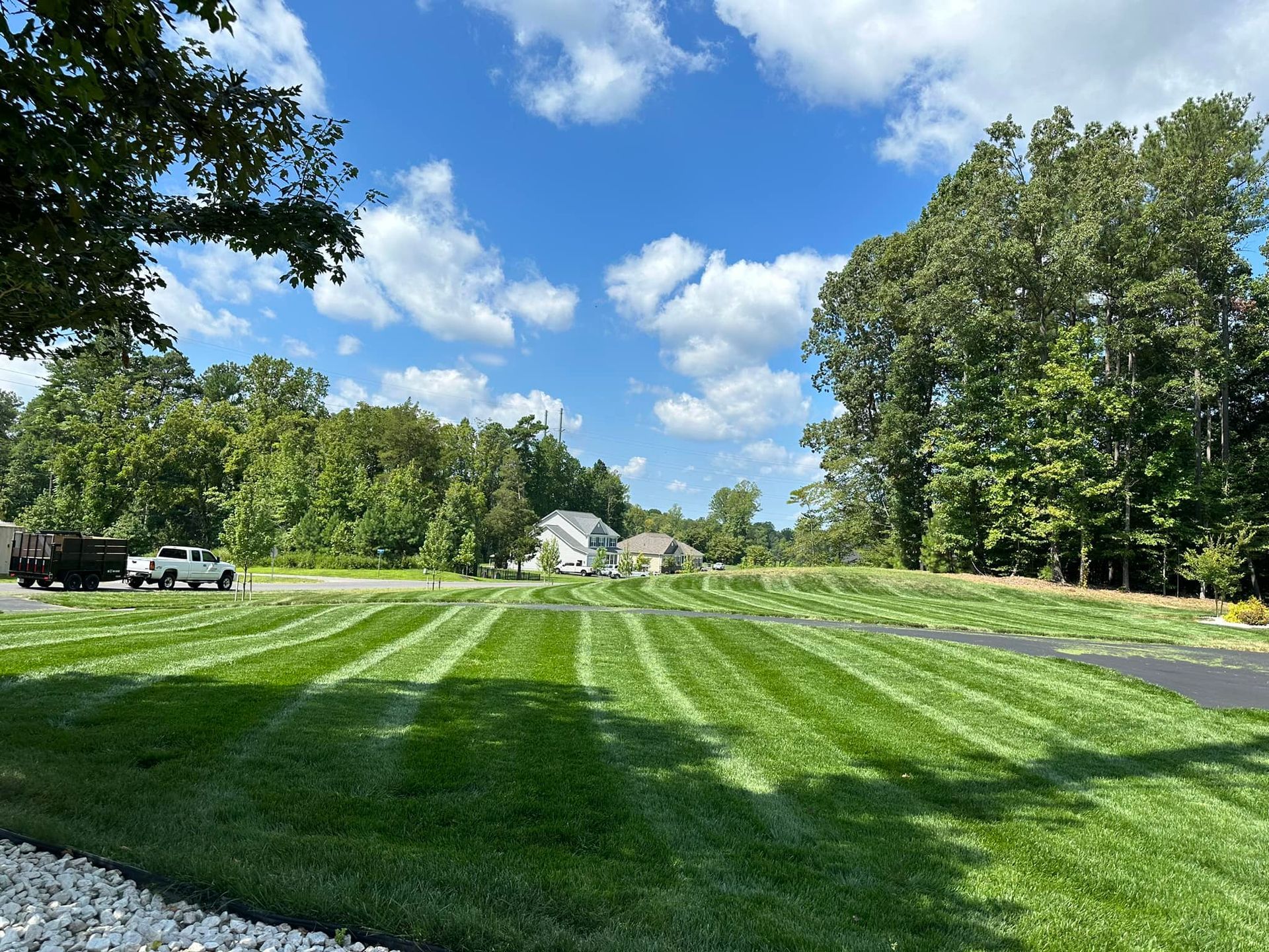 A freshly mowed green lawn with distinct mowing lines, surrounded by trees under a bright blue sky with scattered clouds.