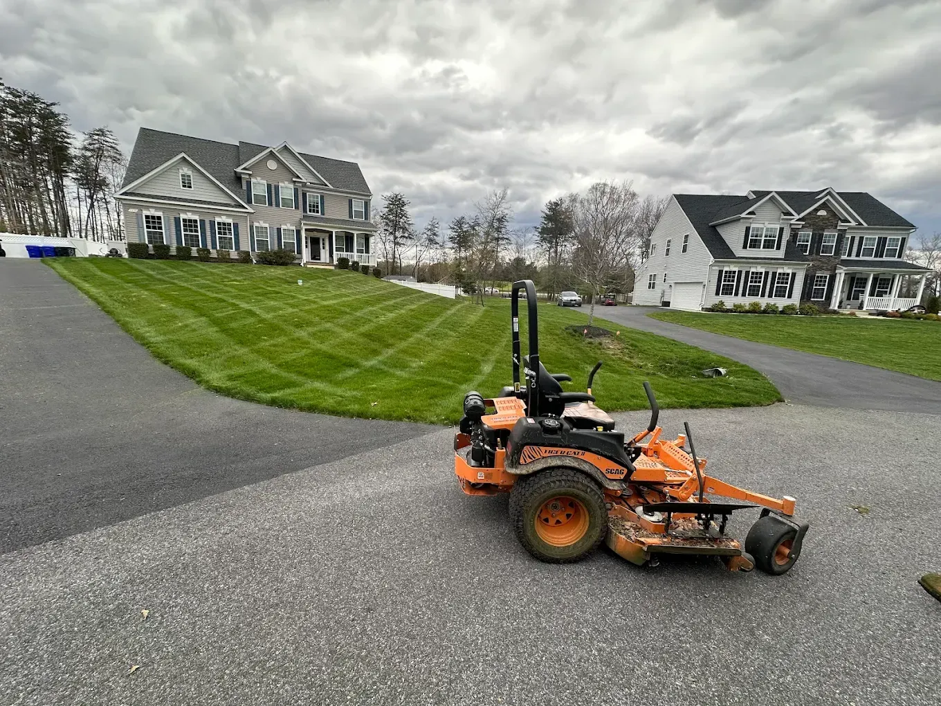 An orange zero-turn mower parked on a gravel driveway in front of a large suburban house with a freshly mown lawn.